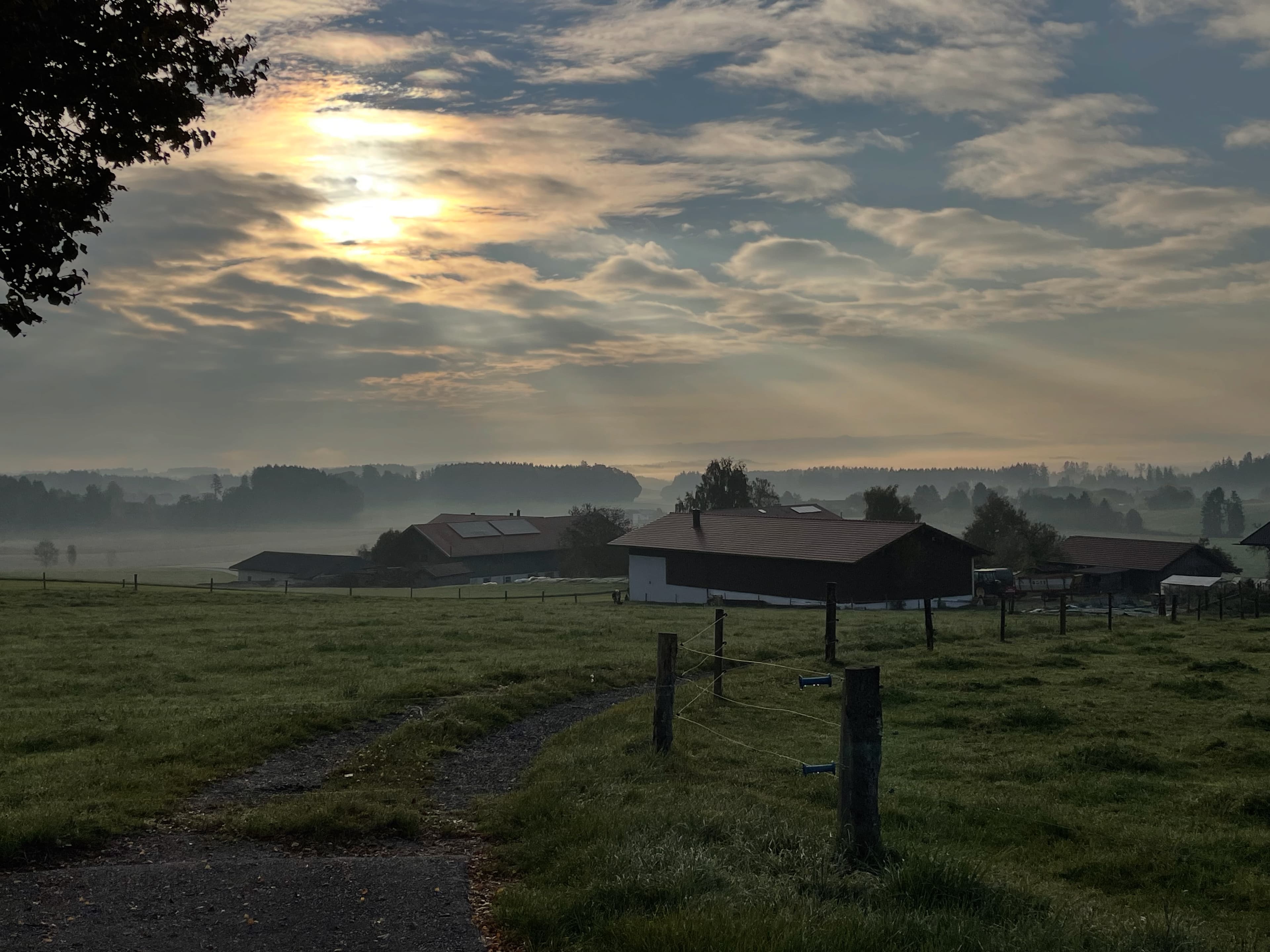 Sunbeams pierce through clouds over a misty countryside with farm buildings and green fields.