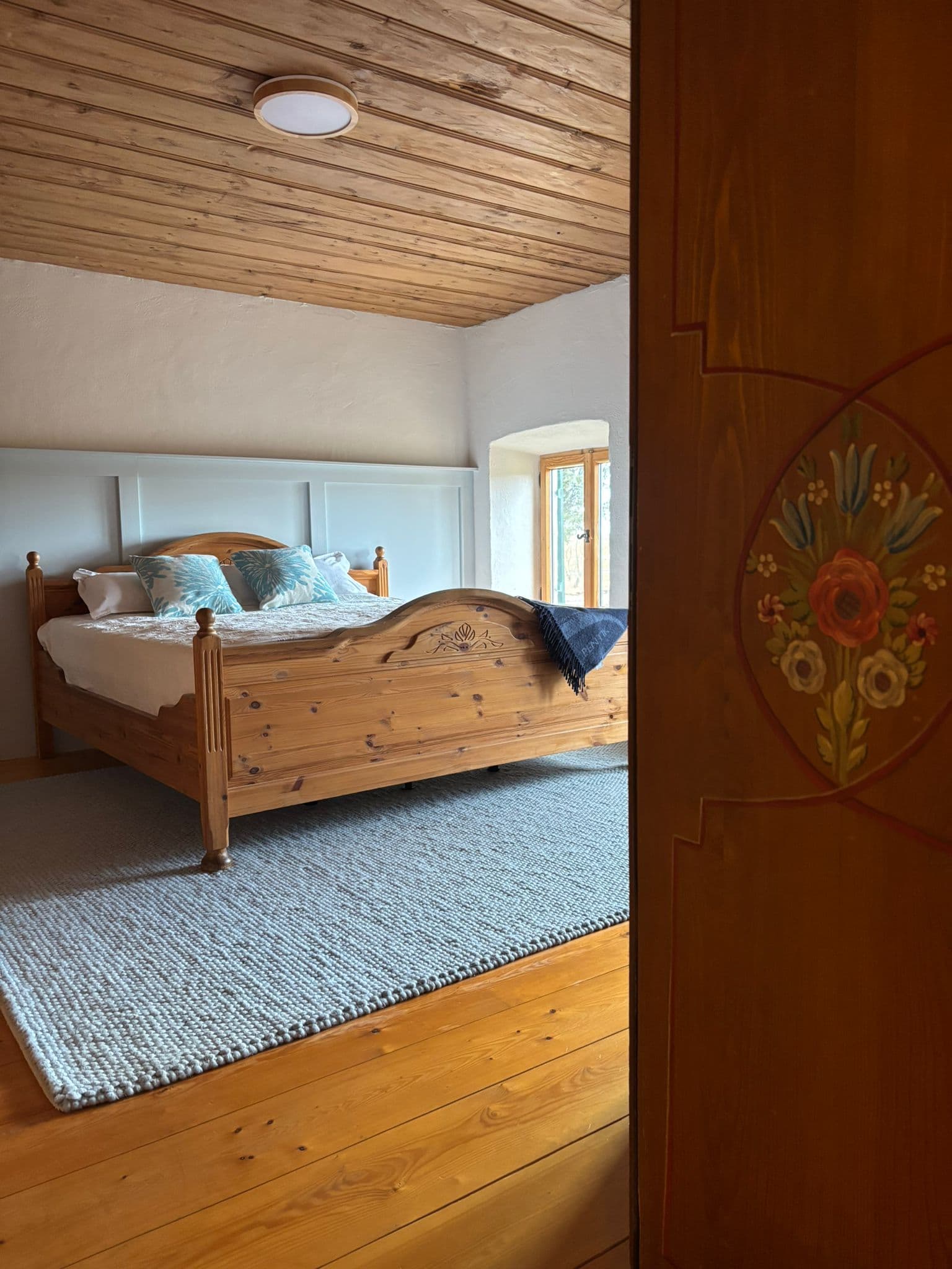 Rustic bedroom with a wooden bed, wooden ceiling, grey rug, and painted floral door.