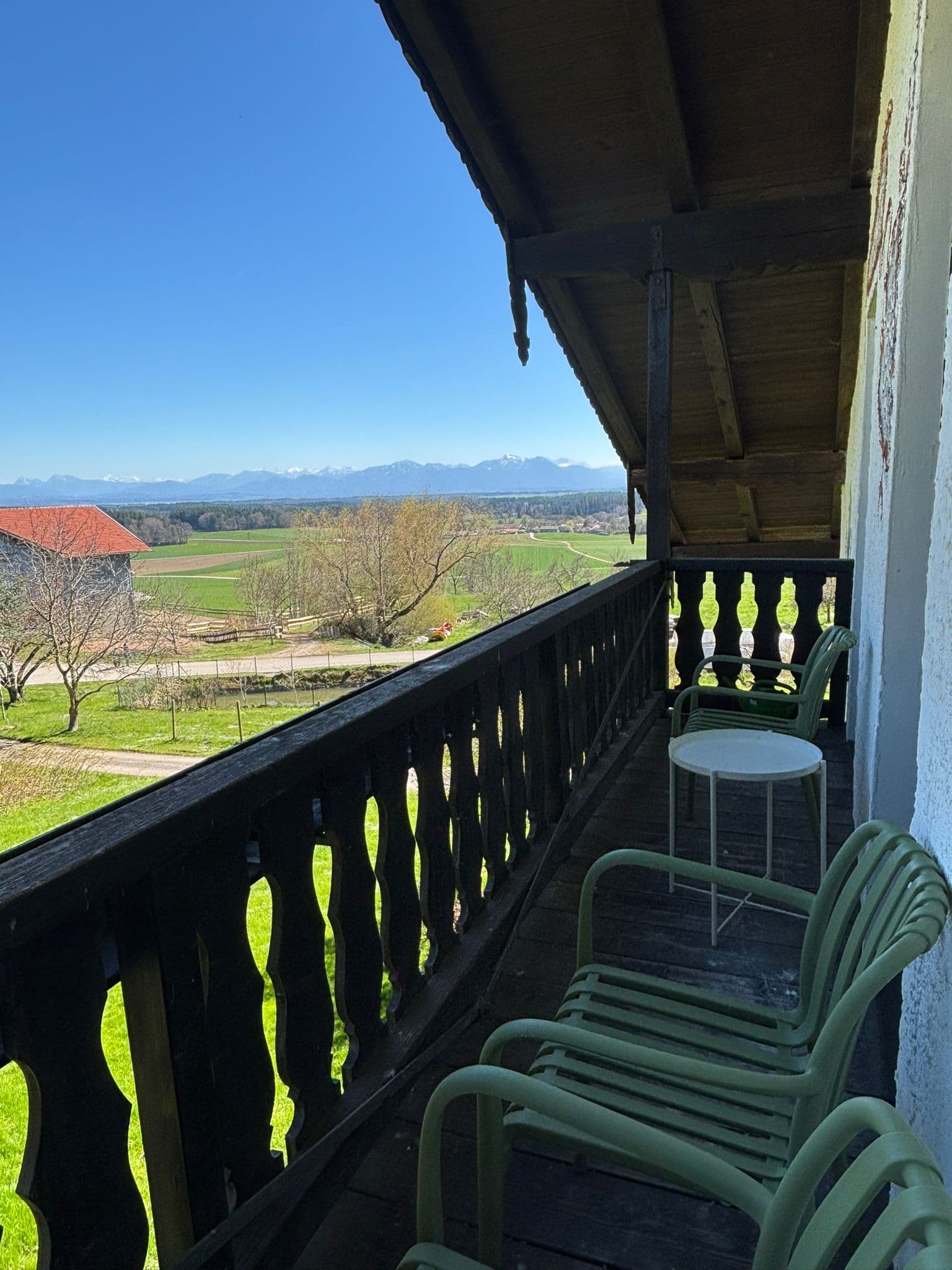 Wooden balcony with green chairs overlooking green fields and distant snow-capped mountains.