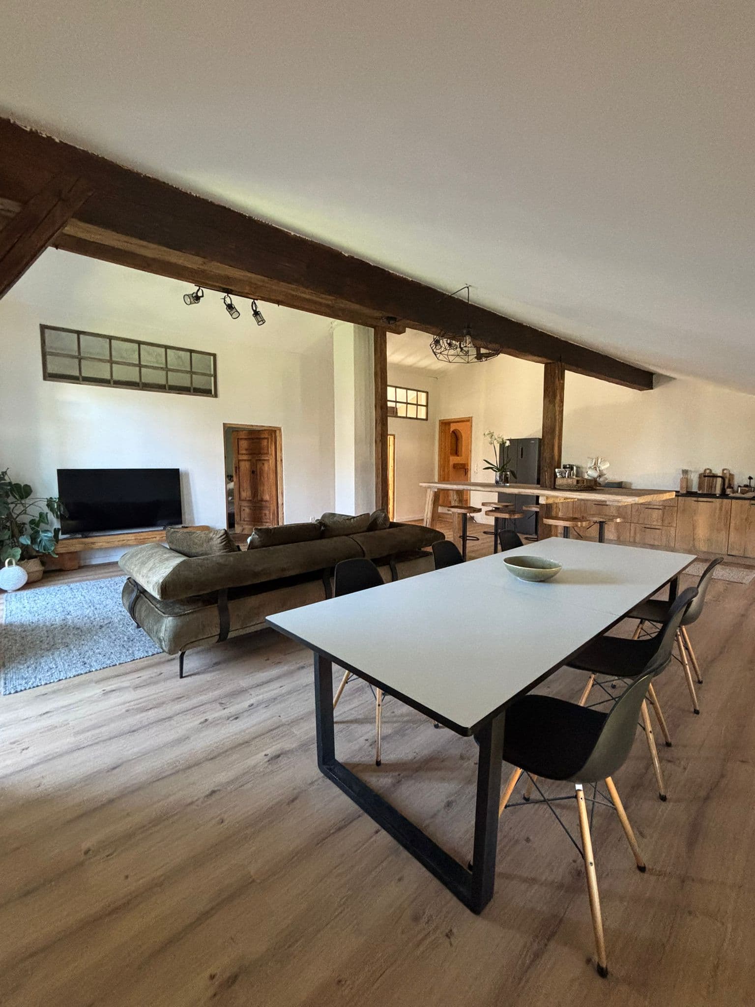 Open-concept living area with a white dining table, green velvet sofa, and rustic wooden beams.