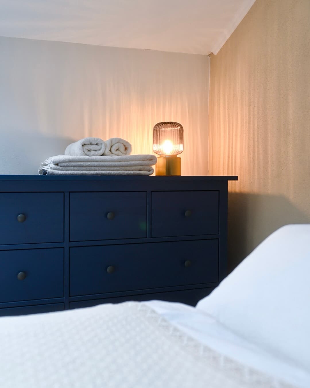 Dark blue dresser topped with folded white towels and a warm glowing ribbed glass lamp.
