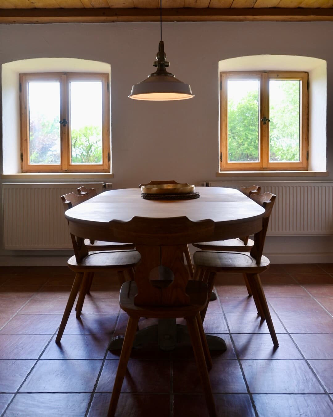 Wooden dining table and chairs under a pendant light between two bright windows.