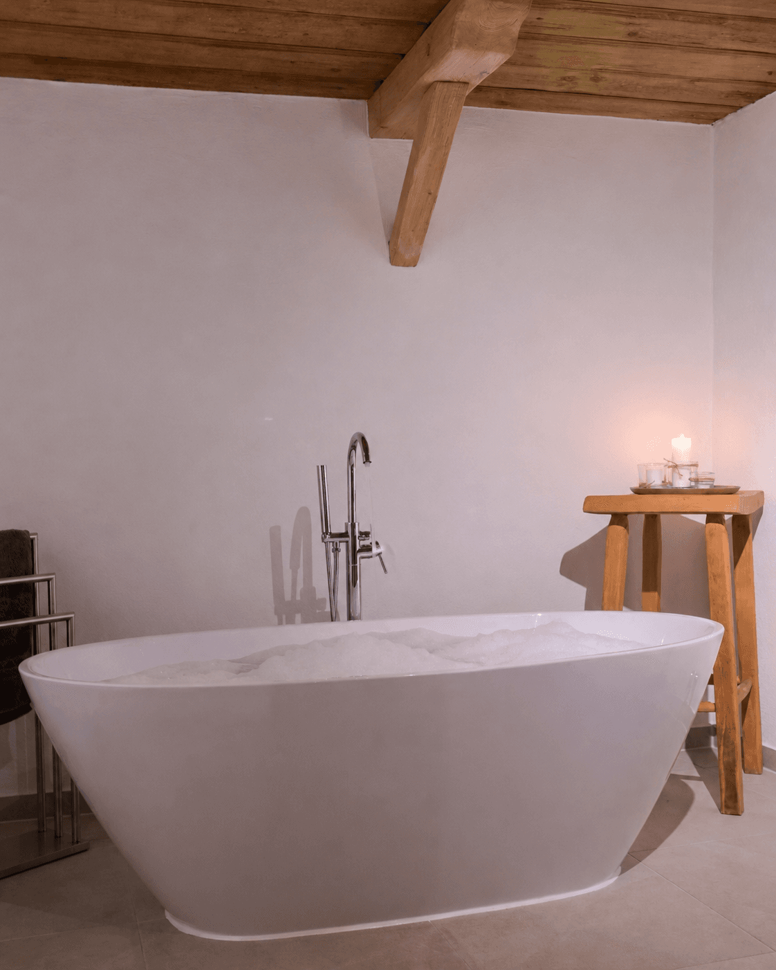 White freestanding bathtub filled with bubbles next to a wooden stool and a lit candle.