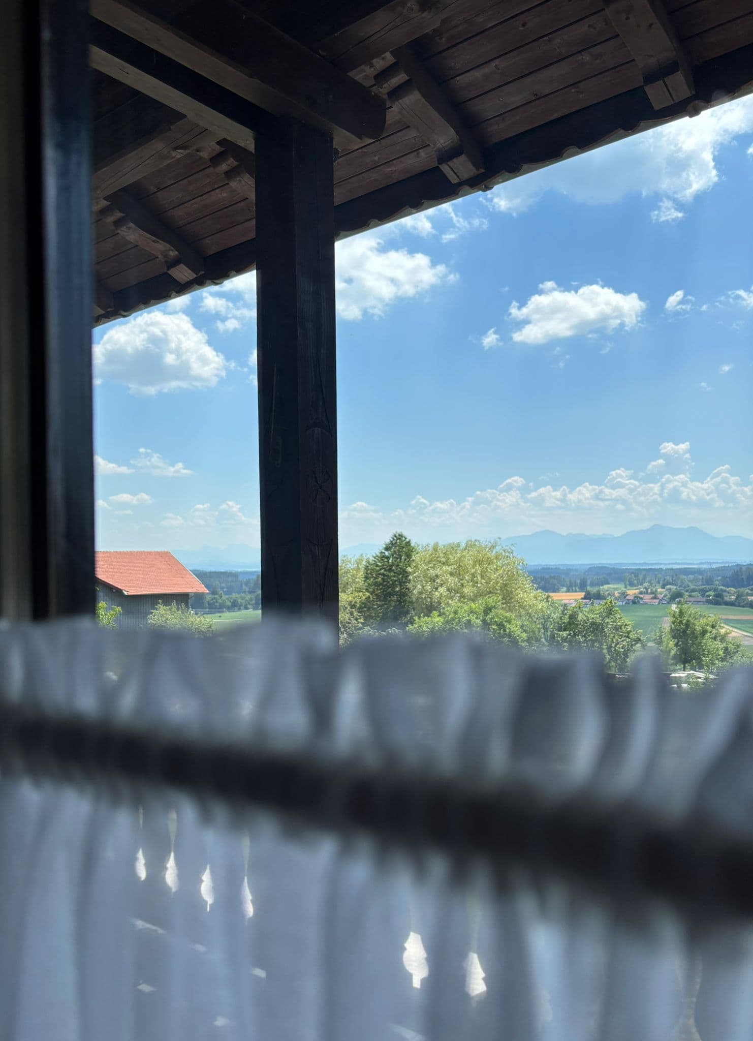 View from a wooden balcony overlooking a red-roofed house, green fields, and distant mountains.