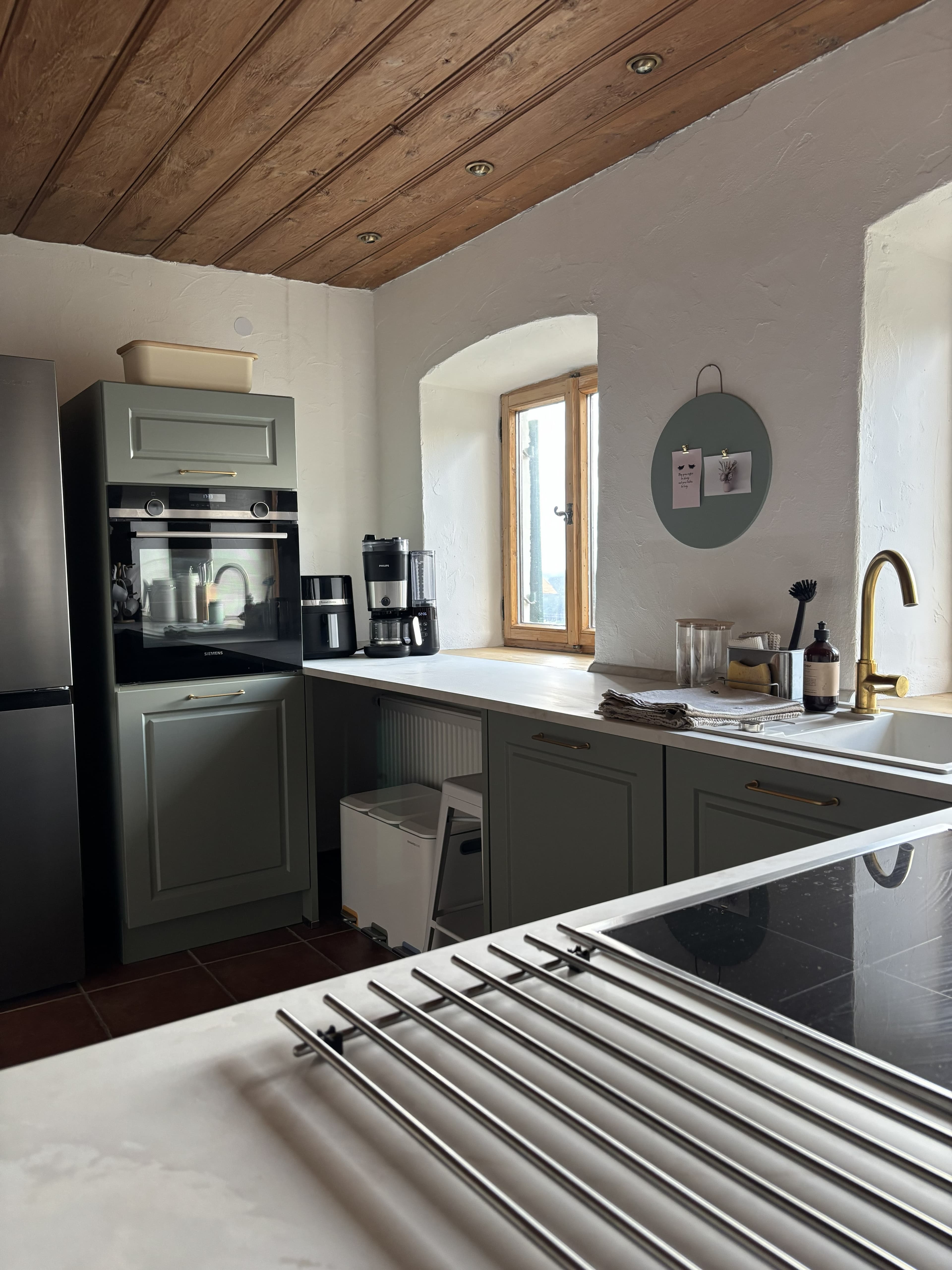 Modern kitchen with sage green cabinets, white countertops, wooden ceiling, and a gold faucet.